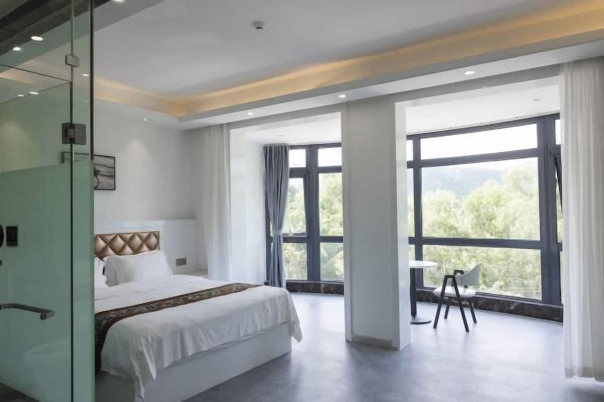 Une chambre d’hôtel moderne avec un grand lit, une petite table et une vue sur la verdure grâce à de grandes fenêtres.