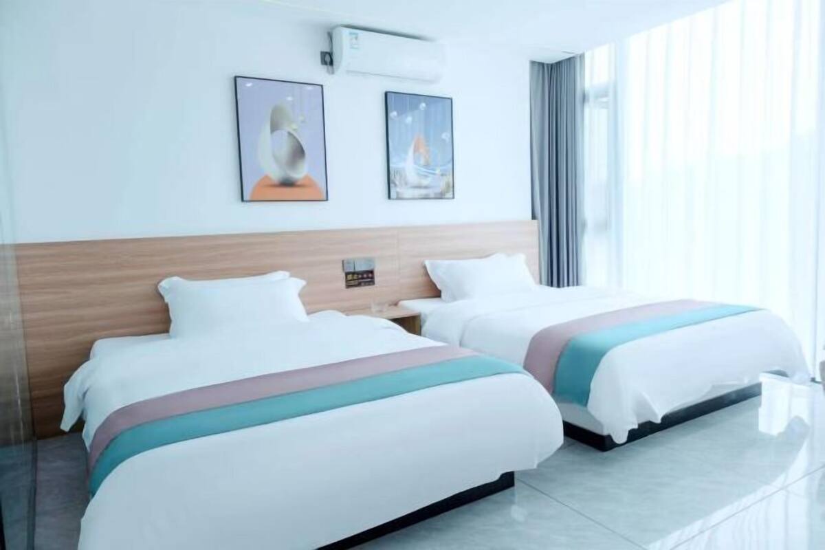 Une chambre d’hôtel avec deux lits, une tête de lit en bois, une fenêtre avec des rideaux et deux œuvres d’art encadrées accrochées au mur.