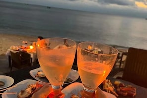 Deux verres d’une boisson claire sont posés sur une table, accompagnés d’assiettes d’huîtres, le tout sur fond de plage.