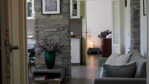 A modern living room with a stone accent wall, a grey sofa, a small table with a green vase, and a kitchen in the background. - Pottery House (Carate Urio)