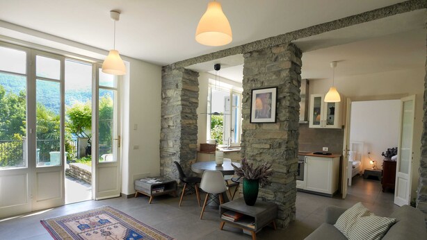 A spacious living area with a stone accent wall, a gray sofa, a dining table with chairs, and a kitchen in the background. - Pottery House (Carate Urio)