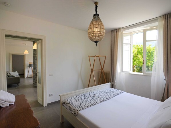A bedroom with a bed, a standing lamp, a window with curtains, and a view of greenery outside.