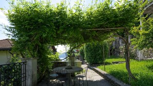 An outdoor patio with a stone table and chairs, surrounded by lush greenery and a view of a body of water. - Pottery House (Carate Urio)