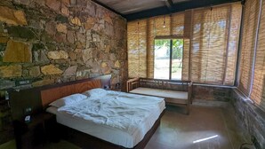 Une pièce avec un mur en pierre, un lit avec une tête de lit en bois, un banc et une fenêtre avec des stores en bambou.