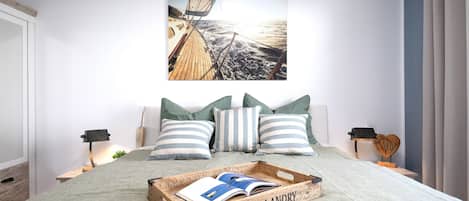 Un dormitorio con cabecera de madera, almohadas a rayas, una mesita de noche de madera con una lámpara, y un cuadro enmarcado de un velero en la pared.