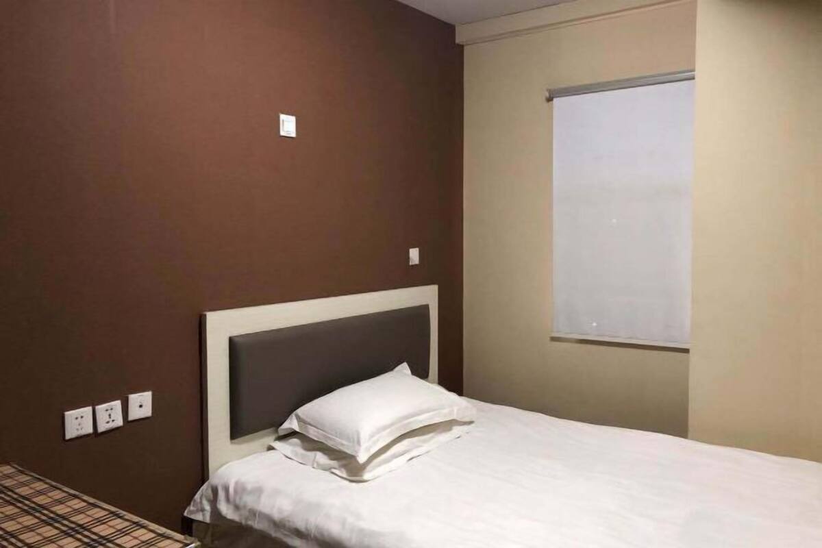 A single bed with a white headboard and pillow in a room with brown walls and a window.