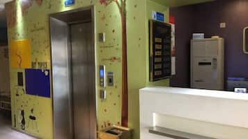 Um elevador com acabamento metálico, um balcão de recepção com bancada branca e uma parede com detalhes decorativos e texto.