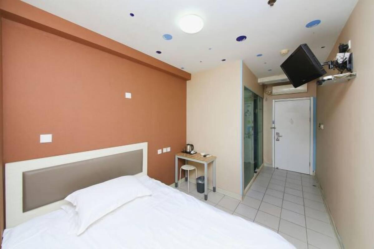 A hotel room with a bed, a small desk with a chair, a television mounted on the wall, and a door leading to another room.