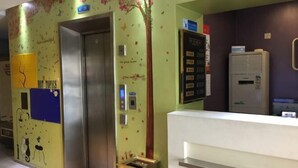 Een lift met een digitaal scherm, een receptiebalie en een prikbord met decoratieve elementen aan de muur.