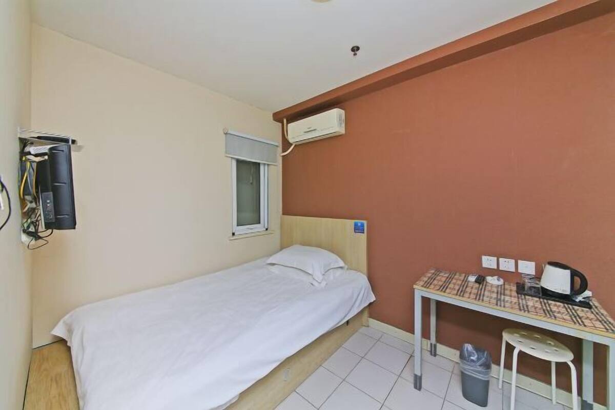 A single bed with a white mattress and pillows, a small table with a patterned tablecloth, a wall-mounted air conditioner, a window with a blind, and a trash bin.