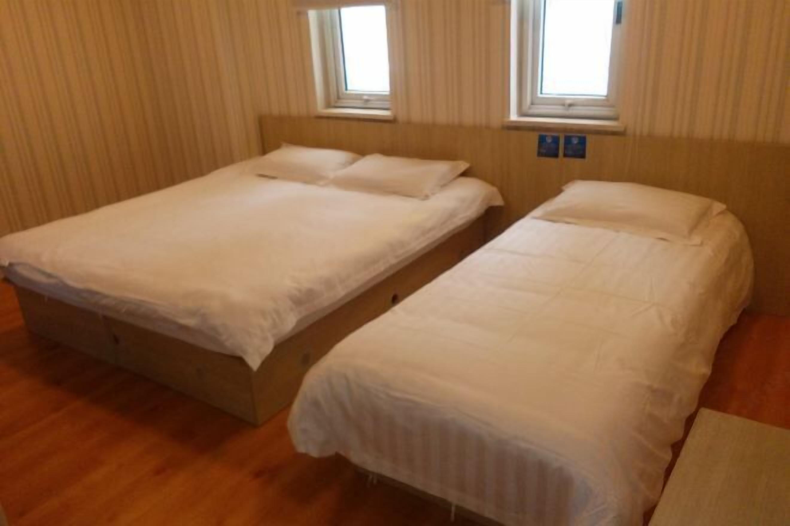 A room with two single beds, wooden flooring, and two windows with curtains.
