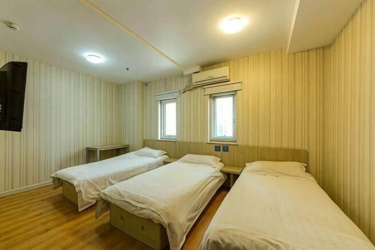 A room with two single beds, a small table, a wall-mounted TV, and an air conditioning unit.