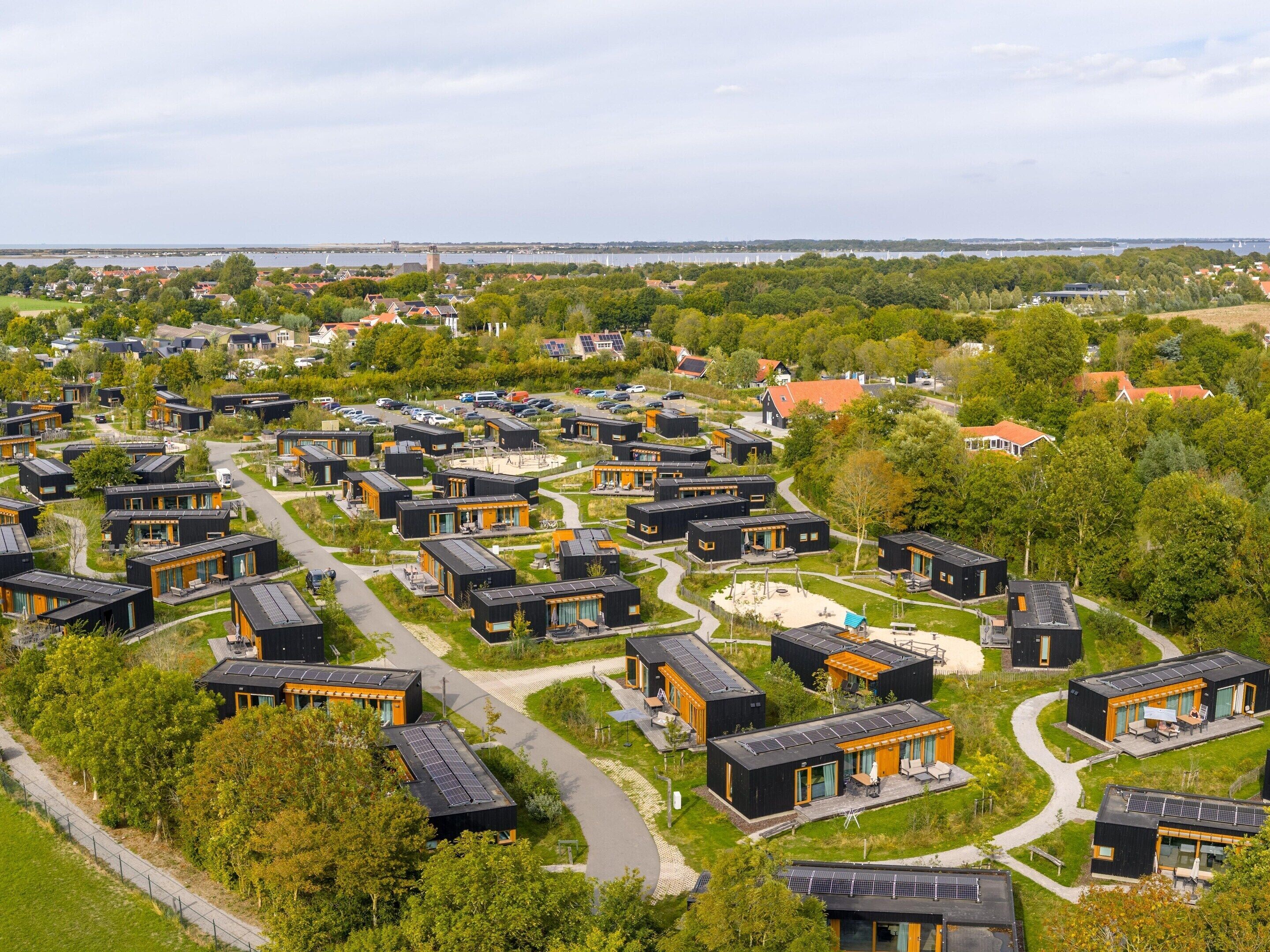 A modern residential area with compact, dark-colored houses arranged in a grid pattern, surrounded by greenery and a body of water in the background.