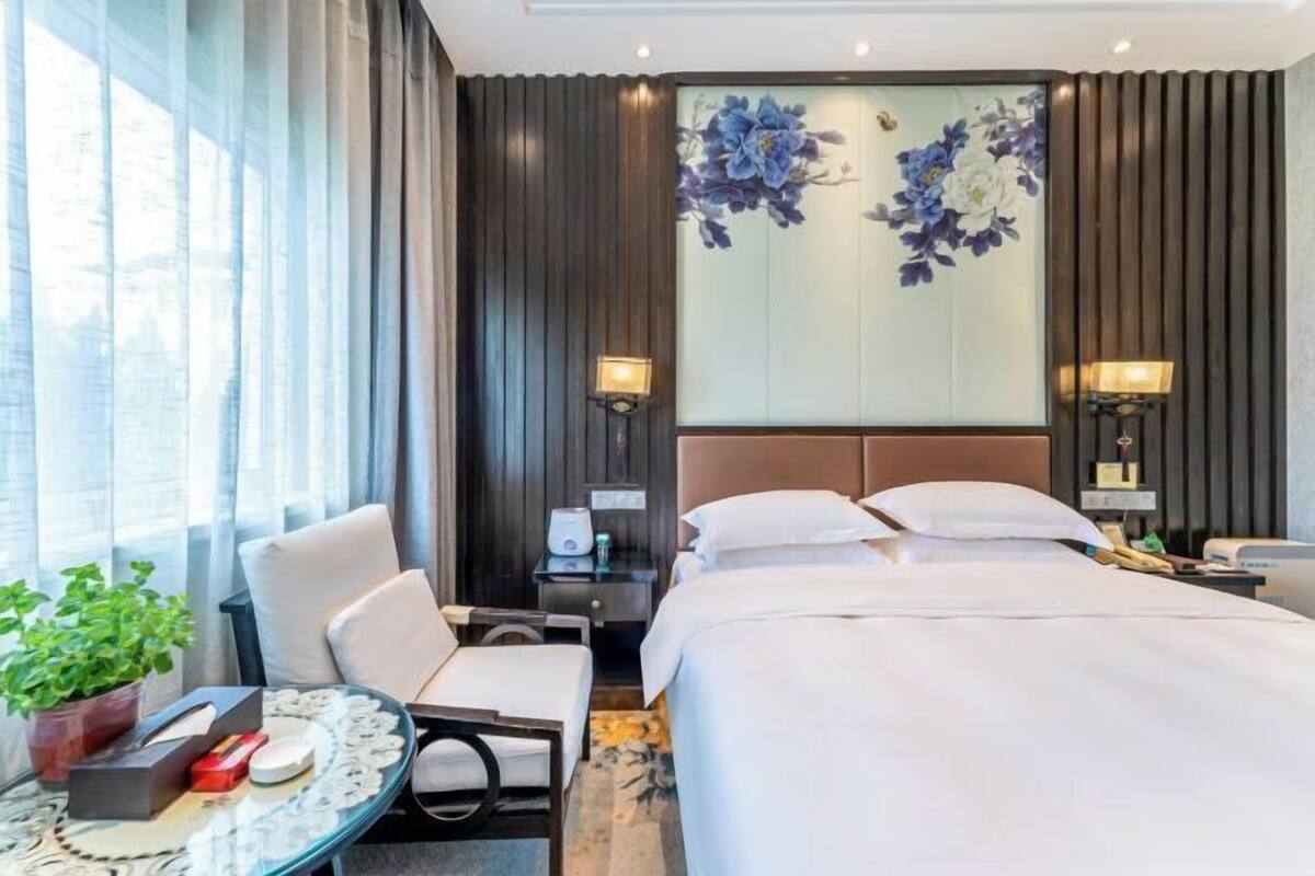 A hotel room with a large bed, a desk with a chair, a small table with a plant, and a wall with a floral mural.