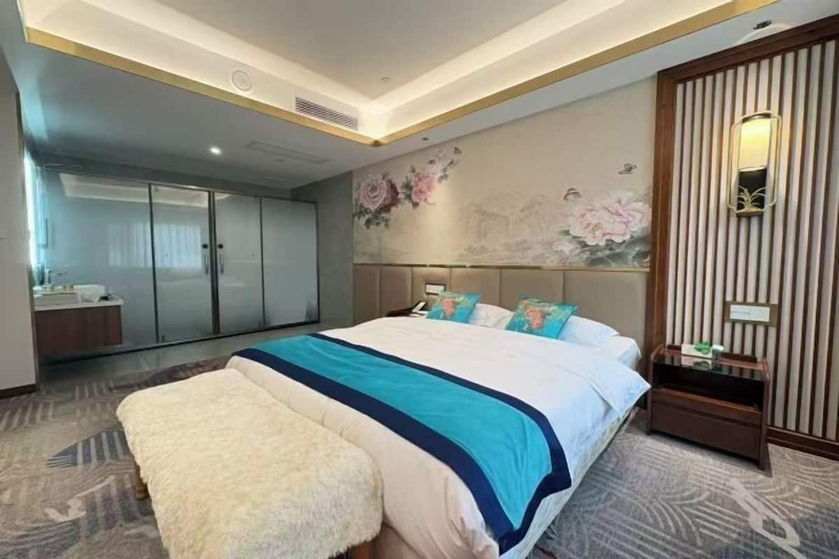 A modern hotel room with a large bed, a fluffy white bench, a nightstand, a mirror, and a sliding door closet.