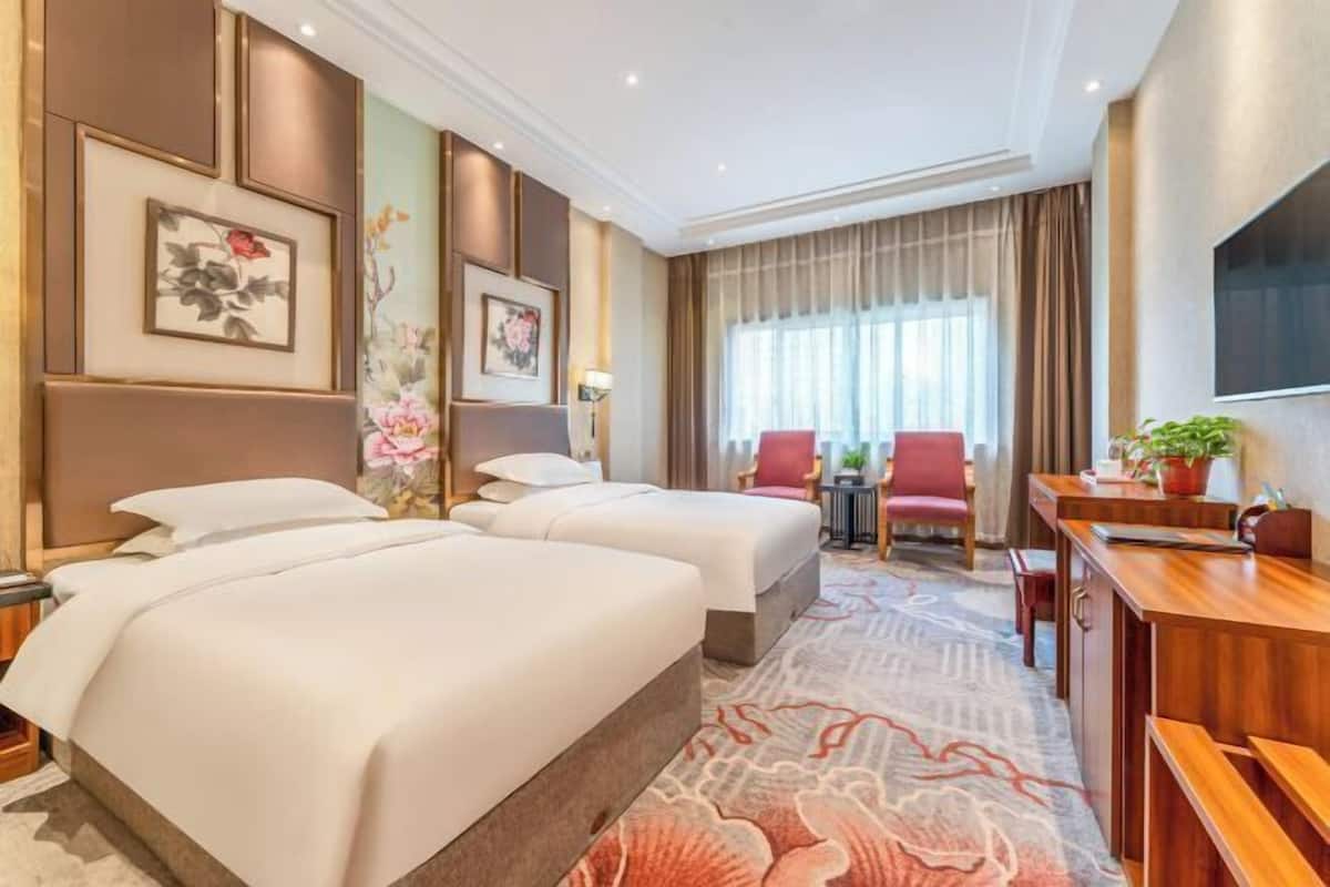 A hotel room with two beds, a desk, a chair, a TV, and a window with curtains.