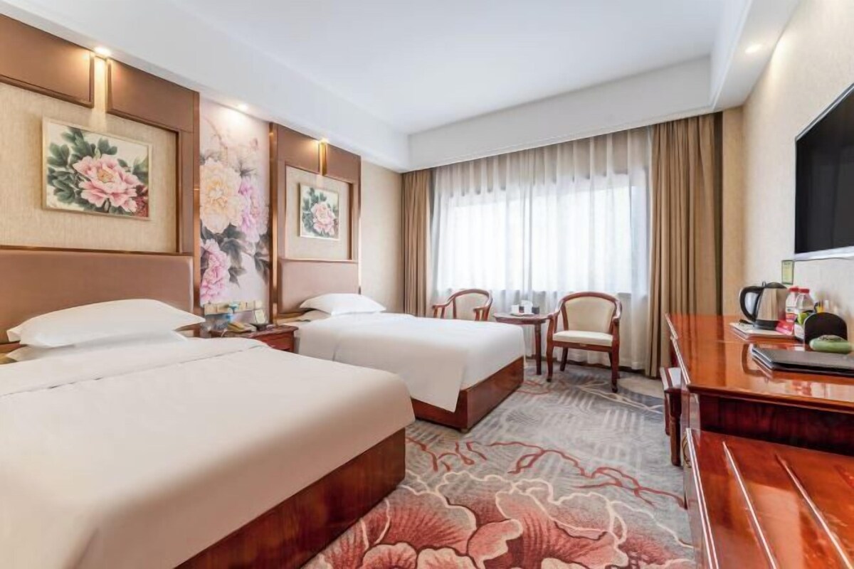 A hotel room with two beds, a desk, a chair, a TV, and a large window with curtains.