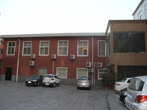 Un estacionamiento con varios autos, un edificio de ladrillo rojo con múltiples ventanas y unidades de aire acondicionado, y un edificio moderno con grandes ventanales.