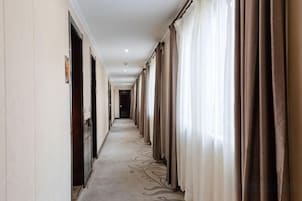 Pasillo de hotel con puertas a ambos lados, alfombra estampada y cortinas a la derecha.