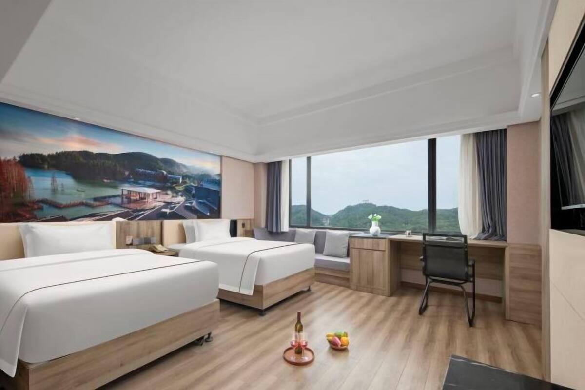 Une chambre d’hôtel avec deux lits, un bureau, une chaise, une grande fenêtre donnant sur un beau paysage et un tableau au mur.