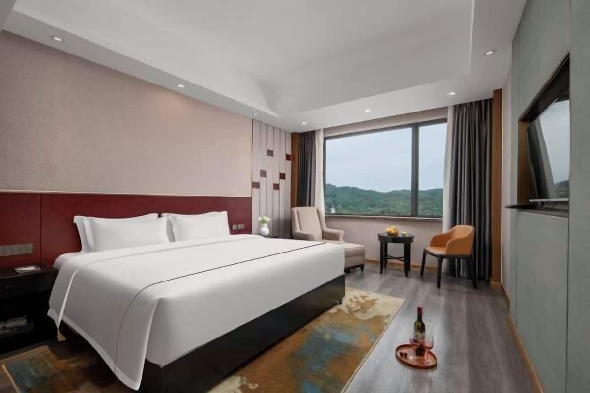 Une chambre d’hôtel avec un grand lit, un bureau, une chaise, une petite table avec une bouteille et un verre, et une vue sur l’extérieur.