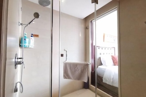 A glass-enclosed shower area with a handheld showerhead, a towel rack, and a glass door leading to a bedroom with a bed and a nightstand. - Shanggao Yingbin Hotel (Shanggao)