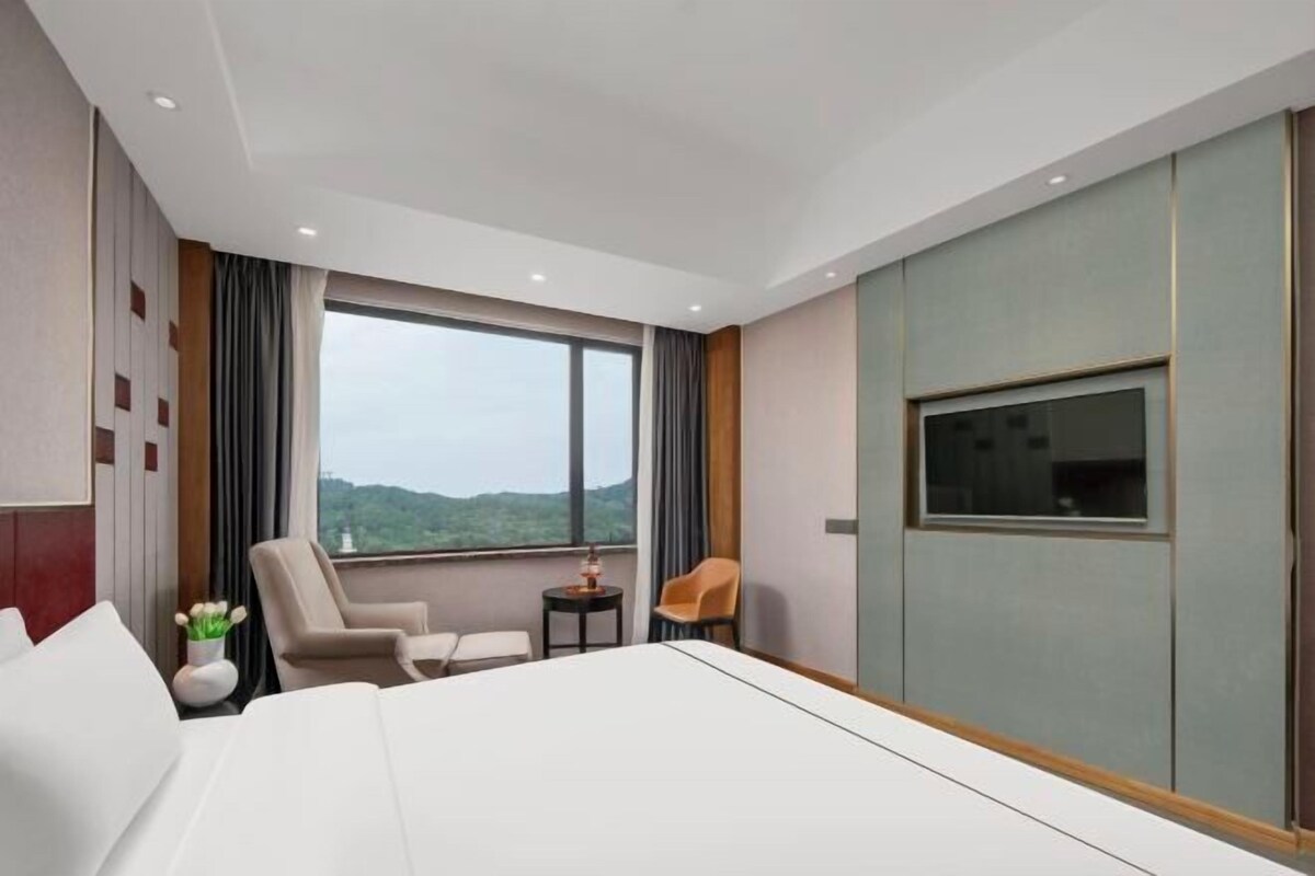 A hotel room with a large bed, a TV, a small table, two chairs, and a view of the outdoors.