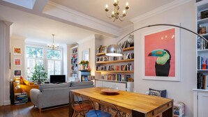 A cozy living room with a wooden dining table, clear chairs, a sofa, and a bookshelf filled with books and framed photos. - Thames-side Retreat, by Veeve (London)