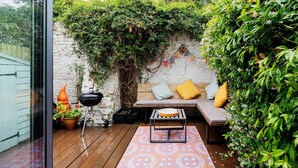 A wooden deck with a bench and a table, surrounded by lush greenery and a brick wall. - Thames-side Retreat, by Veeve (London)
