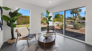 A modern room with a large sliding glass door leading to a landscaped outdoor area. Inside, there is a round wooden table with a woven basket, two wicker chairs, and a large potted plant. - Newly renovated 4-bedroom house with pool and two main suites in Palm Springs.  (Palm Springs)