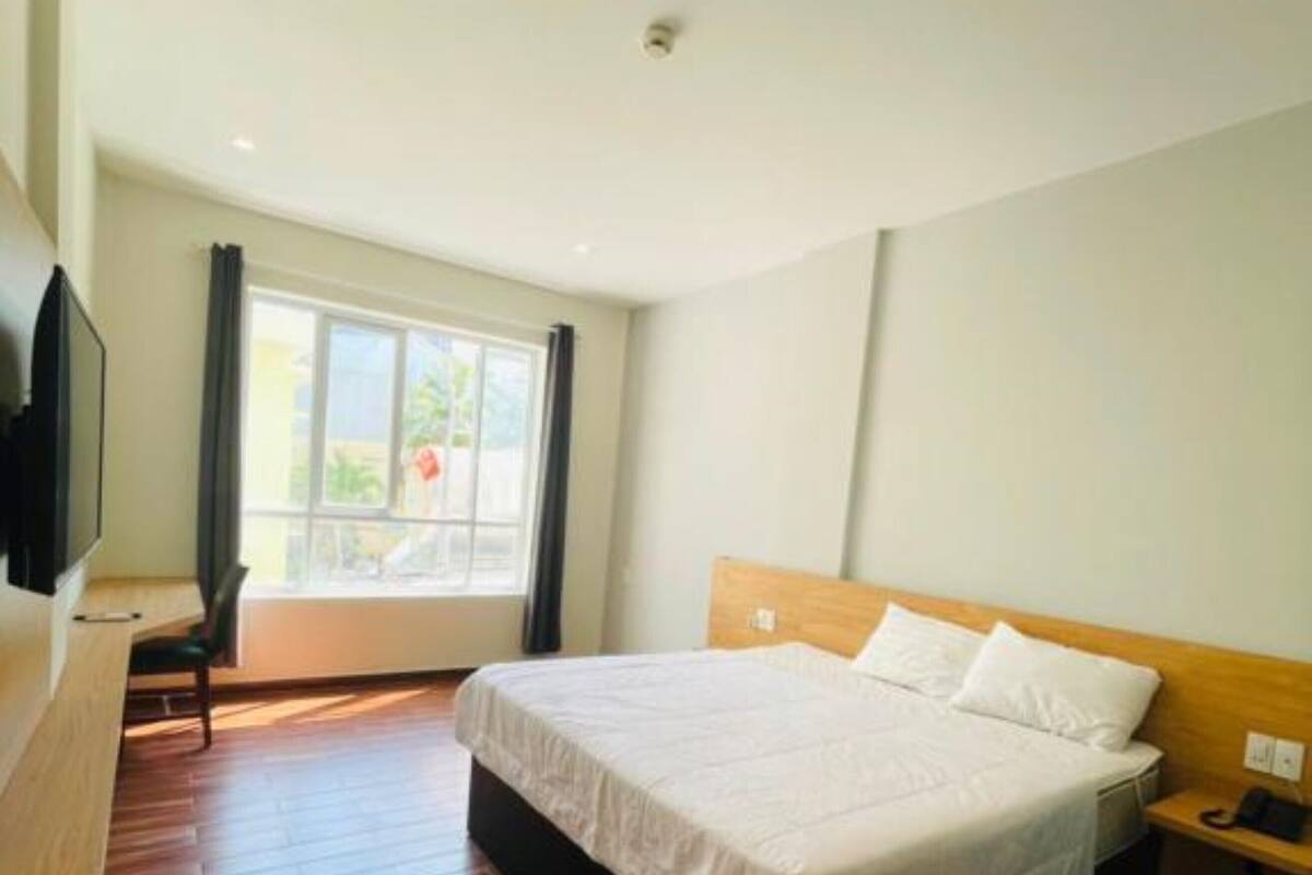 Een hotelkamer met een houten vloer, een bed met witte lakens, een nachtkastje met telefoon en een raam met gordijnen.