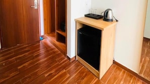 Quarto com soalho de madeira, guarda-roupa de madeira, uma pequena mesa de madeira com uma chaleira, e um eletrodoméstico preto.