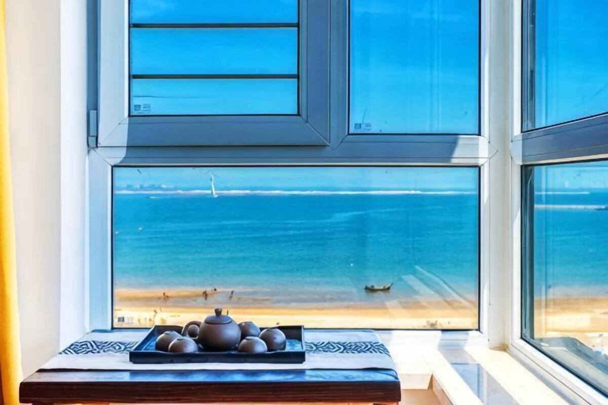Una ventana con vista al mar, una pequeña mesa con una tetera y tazas de té, y un tapete estampado.