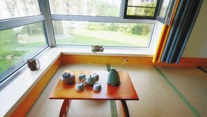 A room with tatami flooring, a wooden table with teapots and a teapot, and a view of a garden through large windows. - Yingkou Vanke Letu Seaview Apartment (Yingkou)