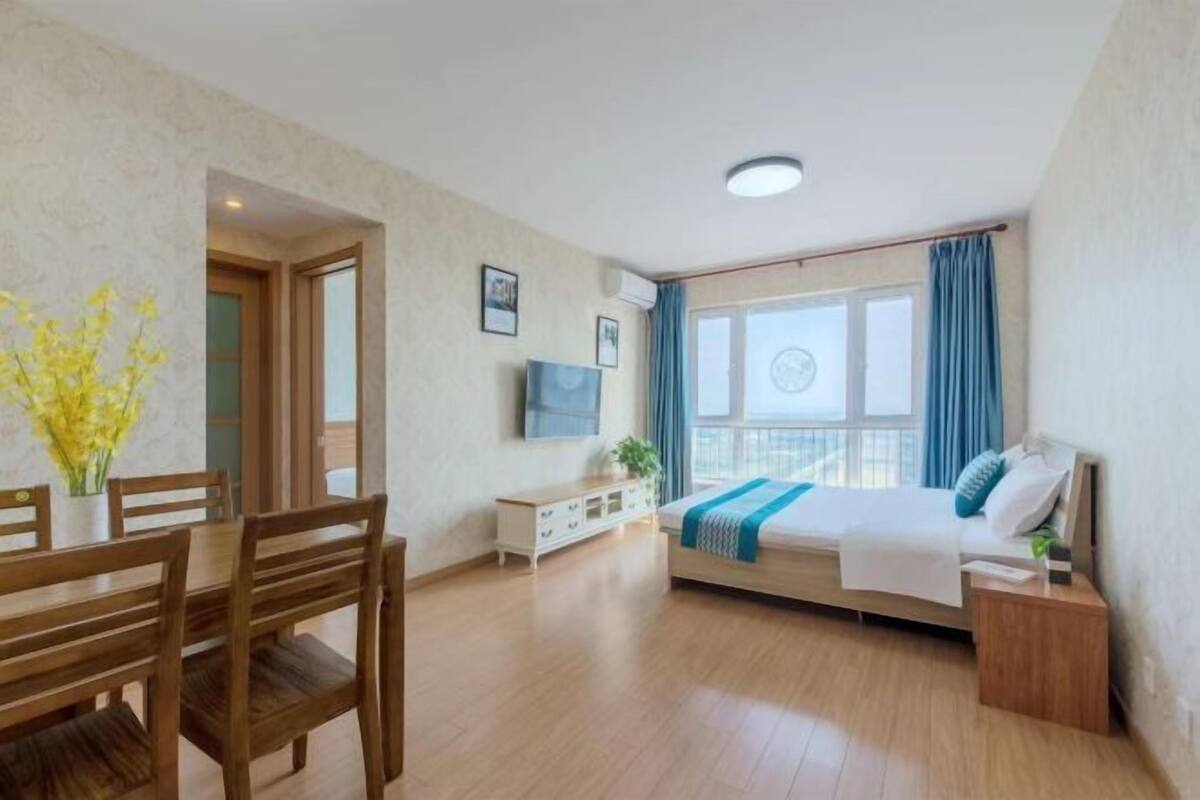 Habitación de hotel moderna con una mesa de comedor de madera, una cama con una manta azul, un televisor en la pared y un balcón con cortinas.
