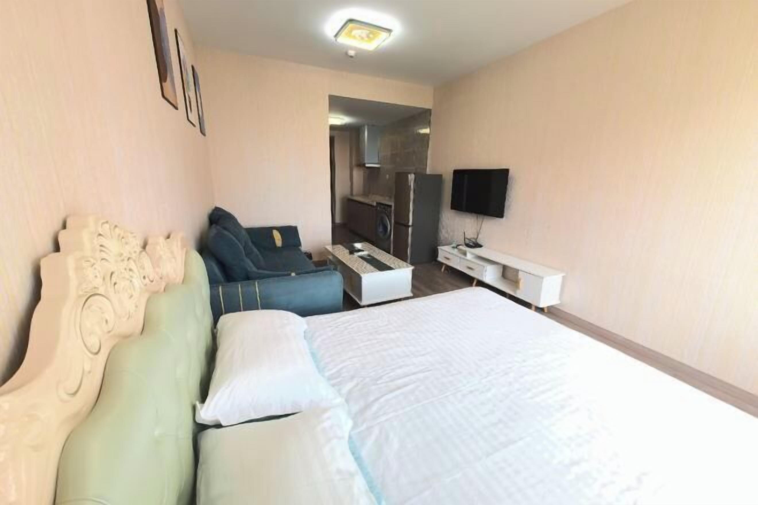 A hotel room with a bed, a sofa, a TV, a desk, and a kitchen area in the background.