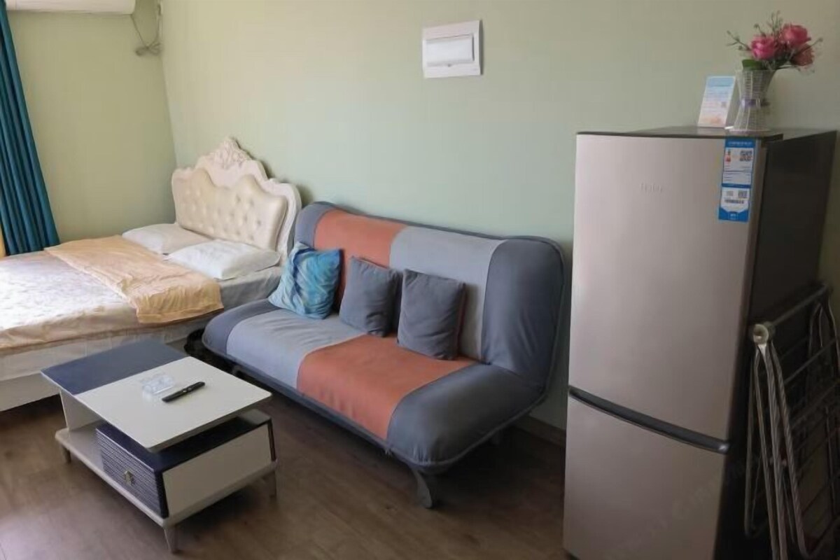 A bedroom with a bed, a sofa, a small table, and a refrigerator.