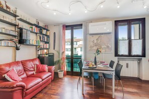 A cozy living room with a red leather sofa, a dining table with chairs, a bookshelf filled with books, and a large window with a view of buildings. - Angera Relax & Pool (Angera)