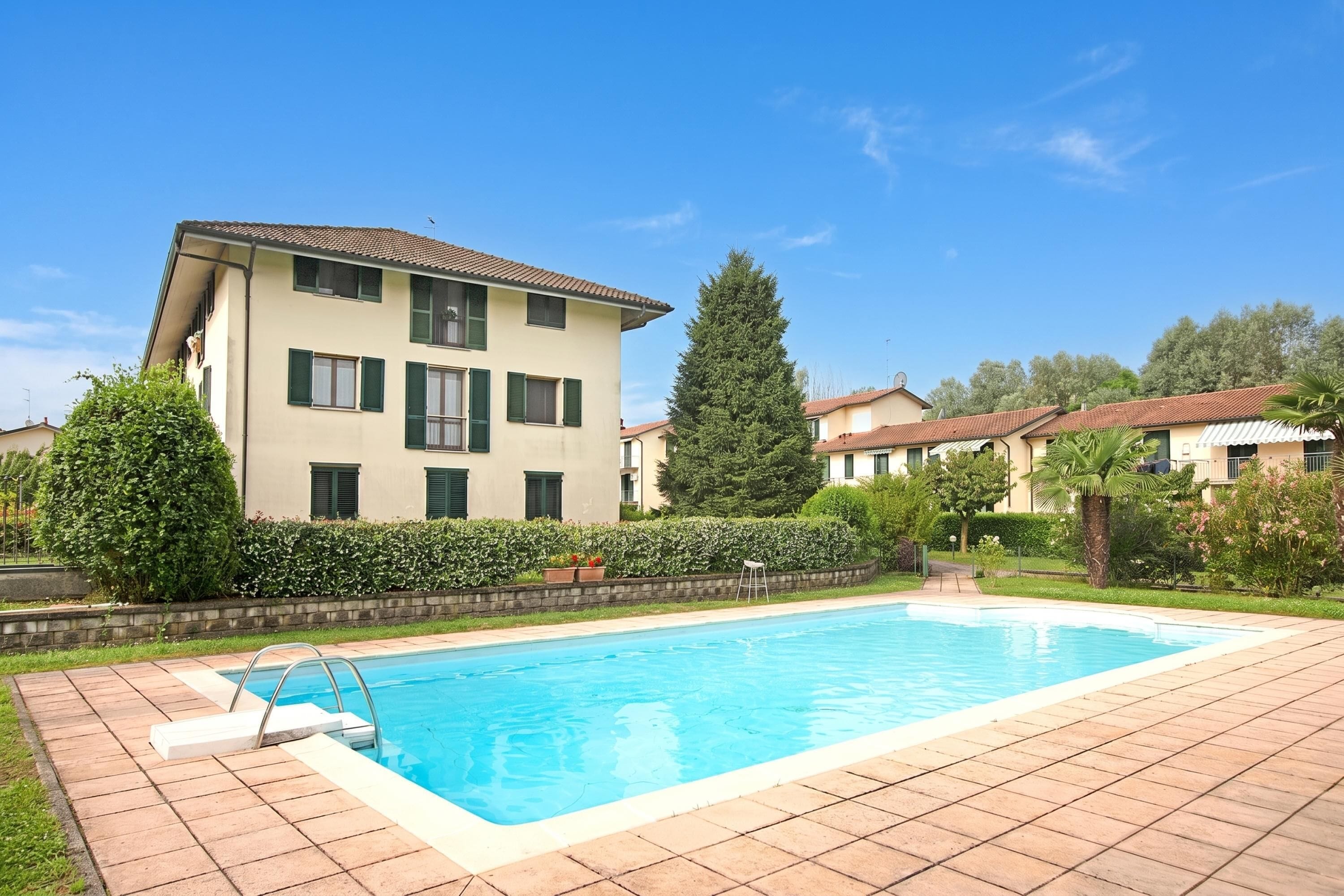 A swimming pool with a tiled deck, a multi-story building with a tiled roof, and a well-maintained hedge.