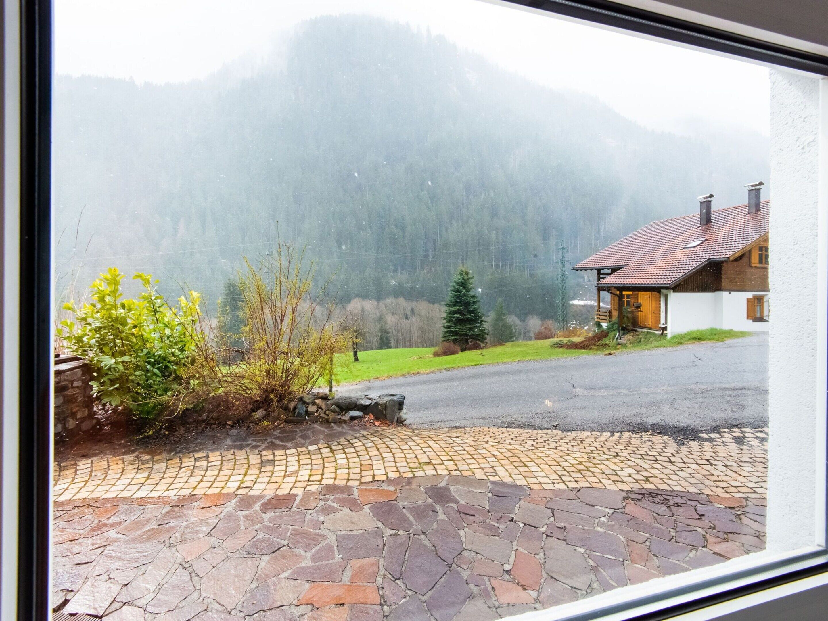 A stone-paved driveway leading to a house with a red-tiled roof, surrounded by greenery and mountains in the background.