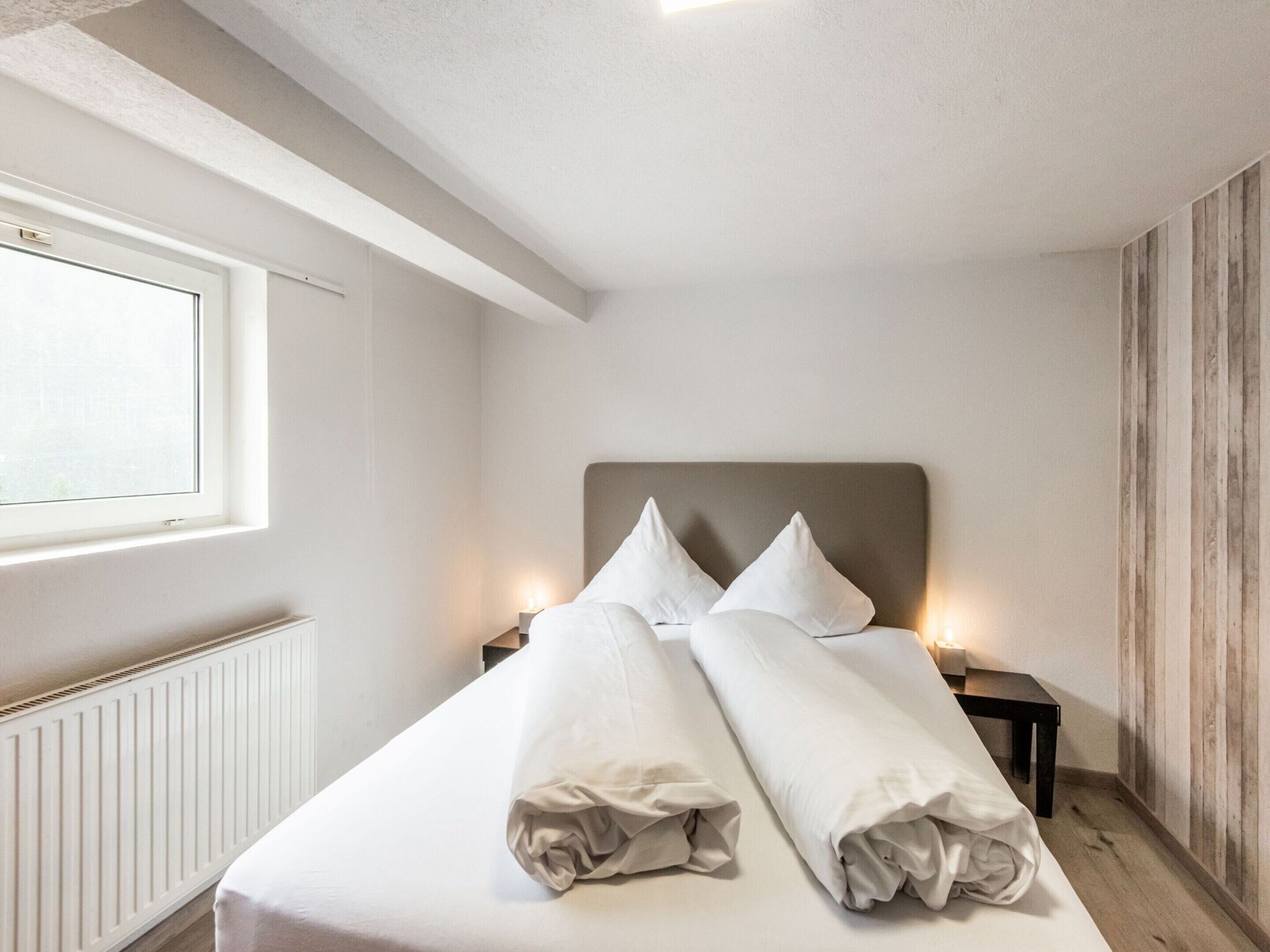A neatly made bed with pillows, a bedside table with a lamp, and a window with blinds.