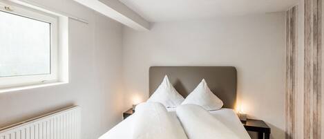 A neatly made bed with pillows, a bedside table with a lamp, and a window with blinds.