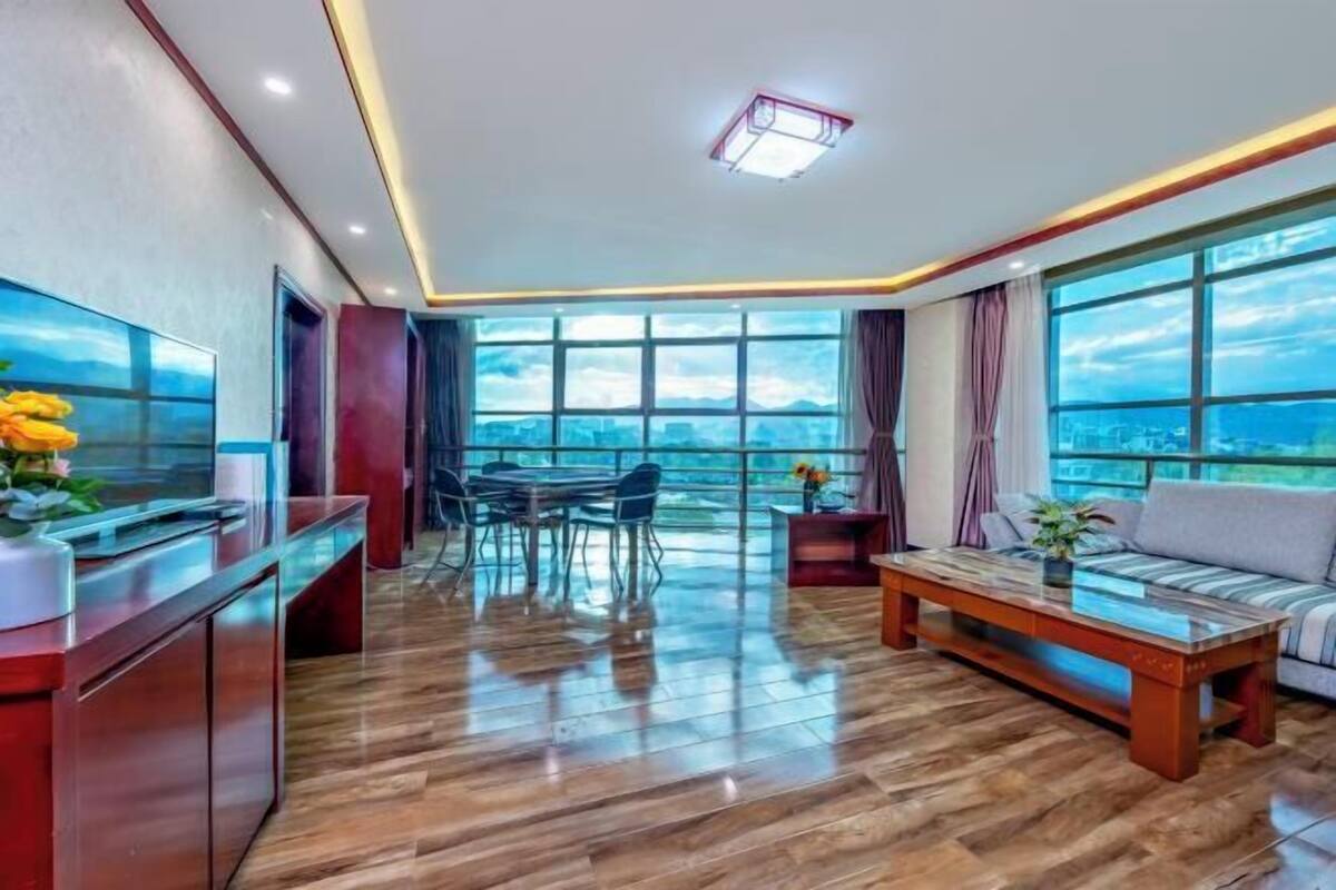 A spacious living area with a large window offering a city view, a wooden coffee table, a sofa, and a TV on a wooden cabinet.