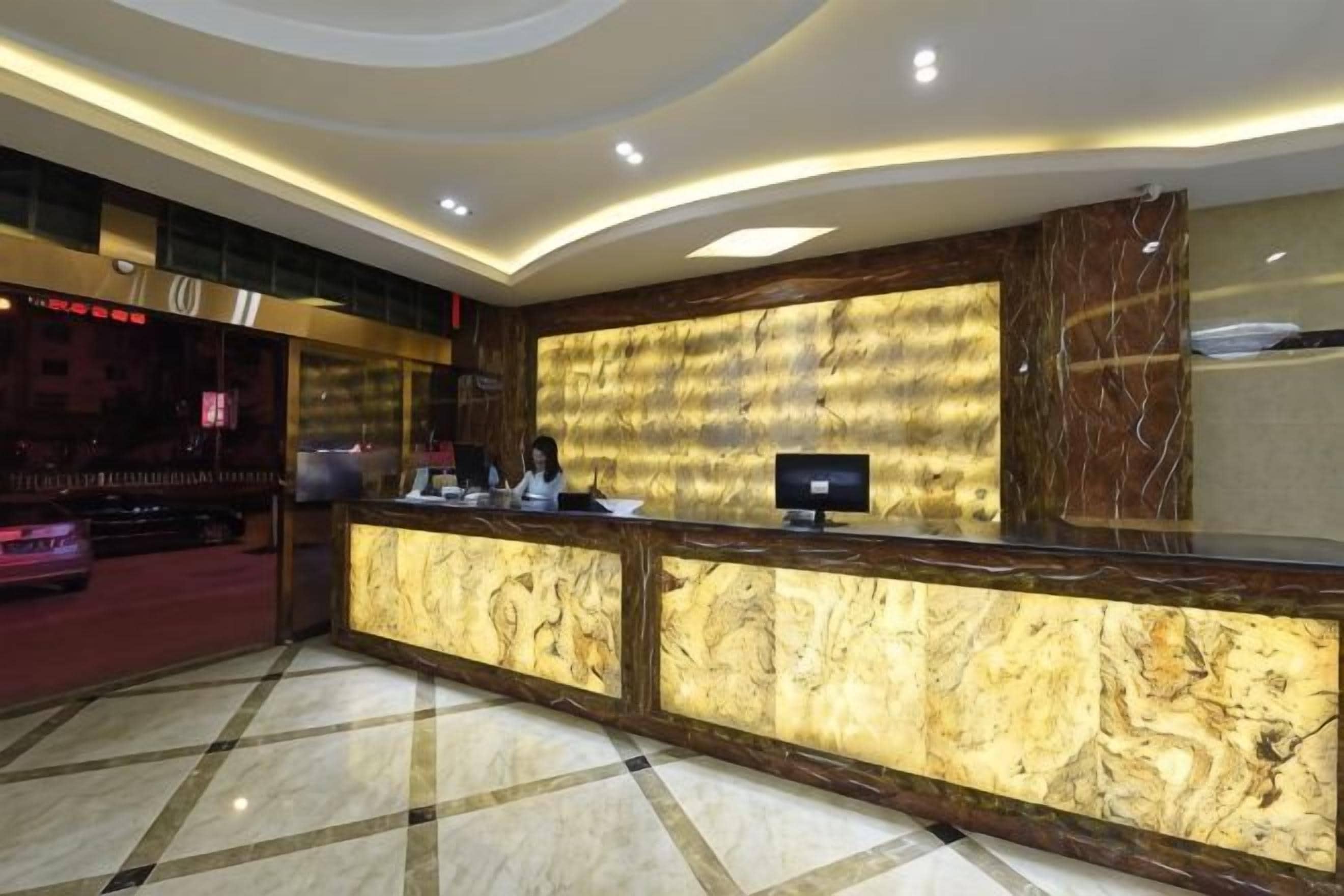 Zona de recepción de un hotel con un mostrador de mármol, un monitor de pantalla plana y una sala de estar con alfombra roja.