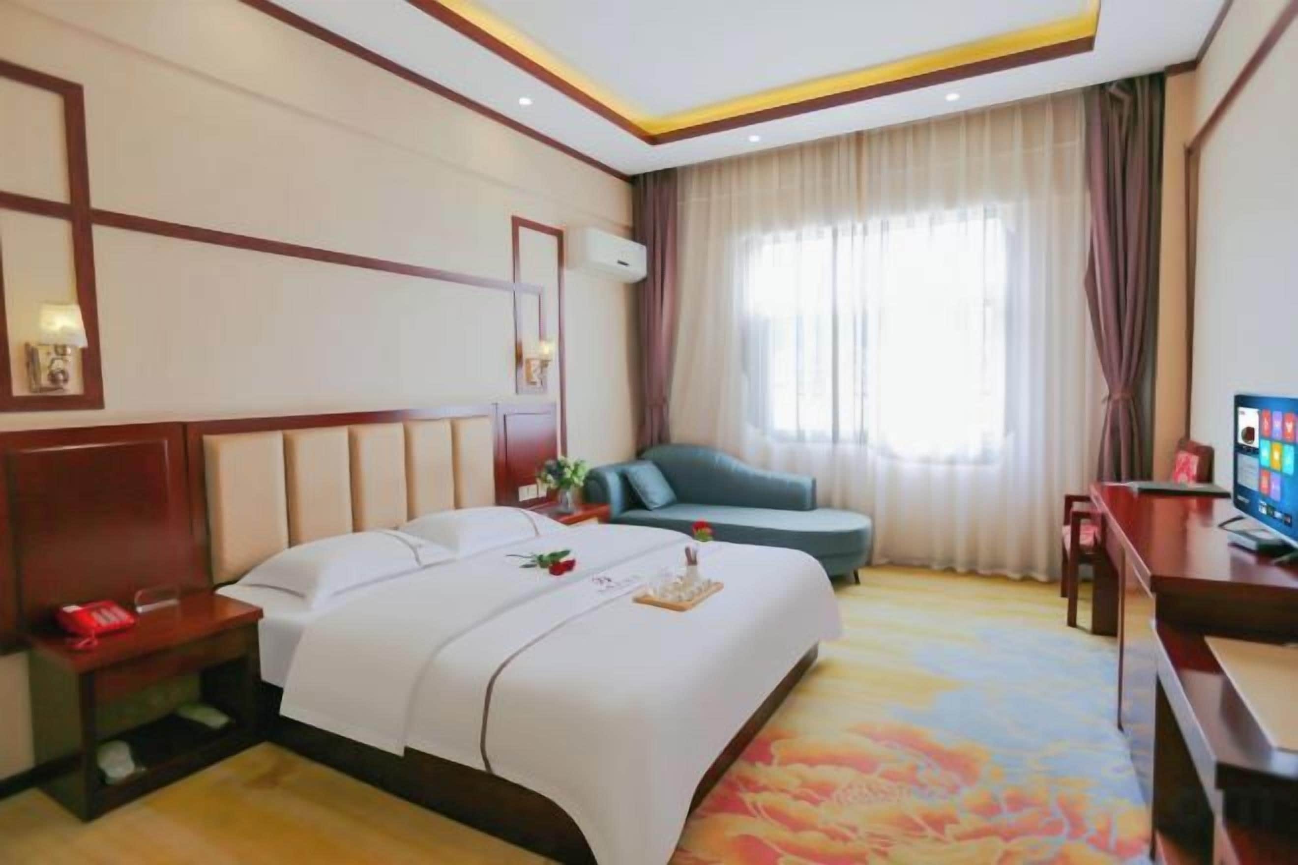 A hotel room with a large bed, a sofa, a desk with a TV, and a window with curtains.