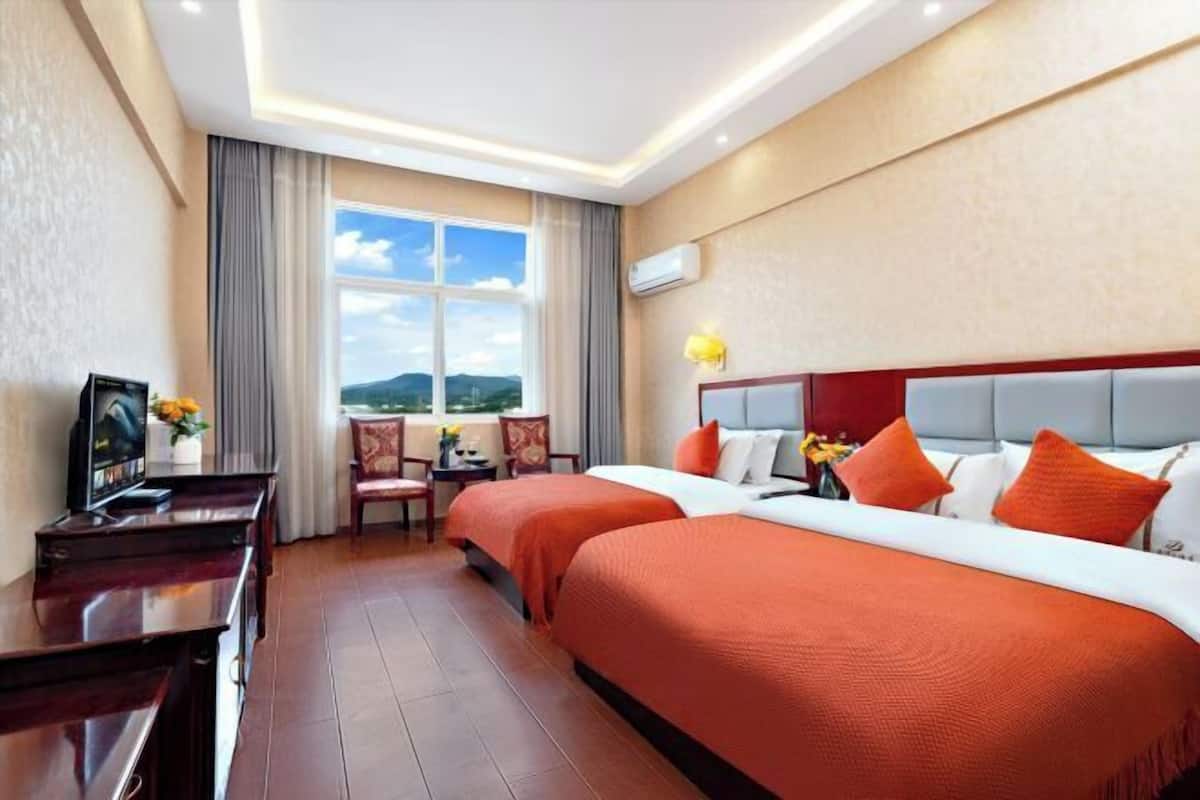 A hotel room with two beds, a desk with a TV, a chair, a window with a view, and a lamp.