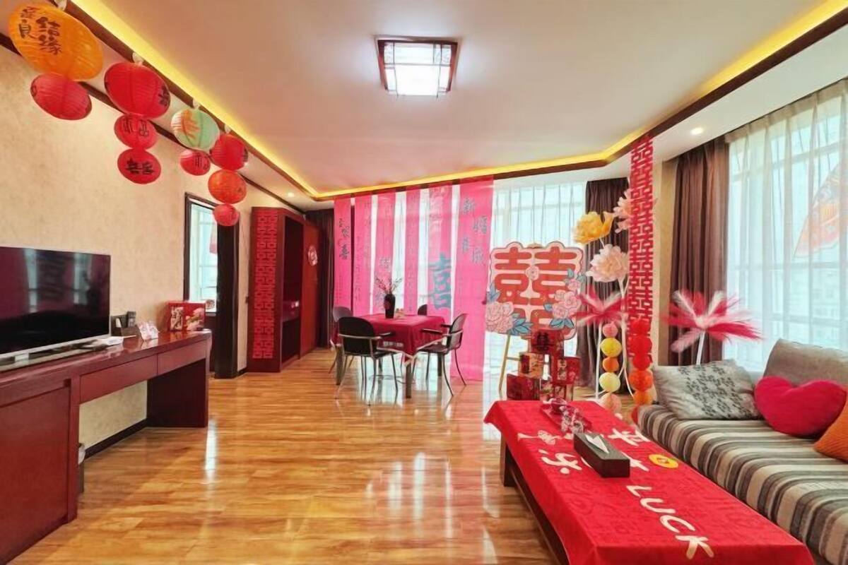 A hotel room with a wooden floor, a TV on a stand, a dining area with a table and chairs, and a sofa with a red tablecloth.