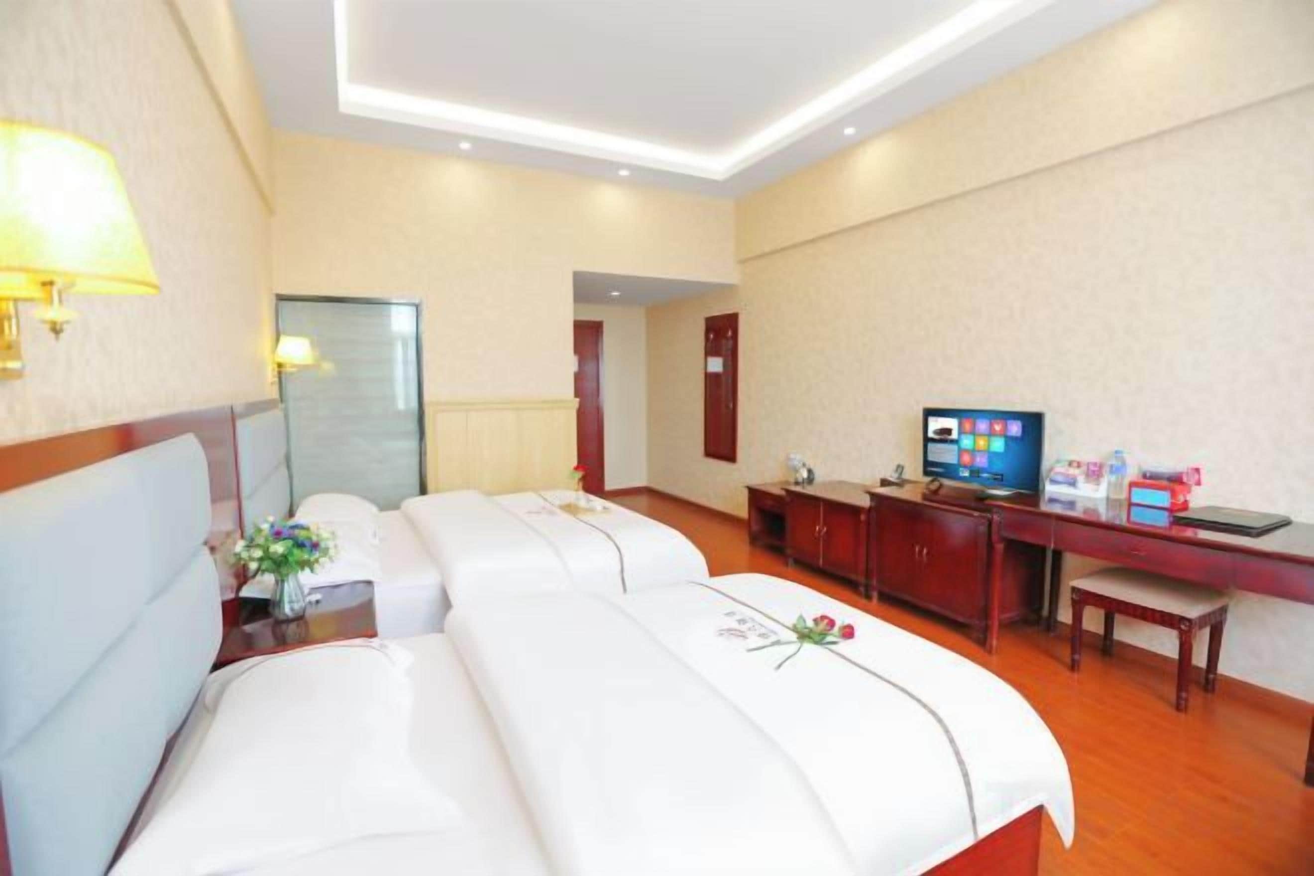 A hotel room with two beds, a desk, a television, and a bathroom visible through a glass door.