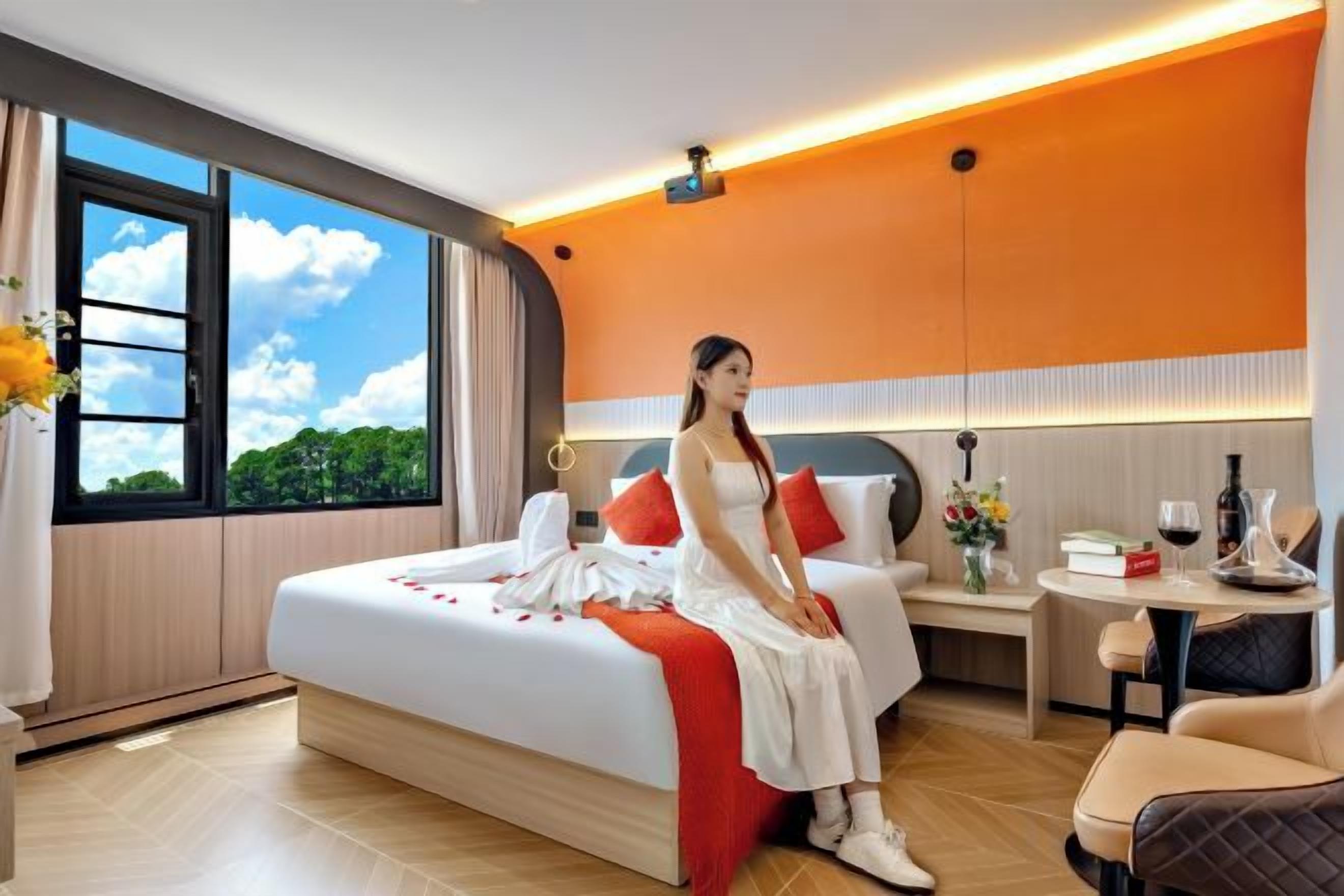 A hotel room with a bed, a bedside table with a lamp, a small table with a vase of flowers, and a view of the sky through a window.