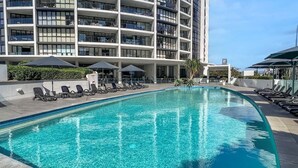 Ein Swimmingpool mit Liegestühlen und Sonnenschirmen, umgeben von einem modernen Gebäude mit mehreren Balkonen.